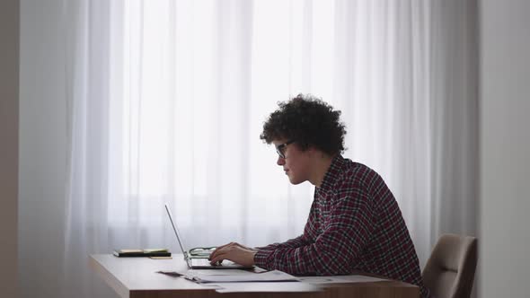 A Curly Man with a Serious Look Works at a Laptop Sitting in a Modern Kitchen