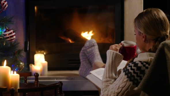 Woman Is Sitting With Cup Of Hot Drink And Book Near The Fireplace