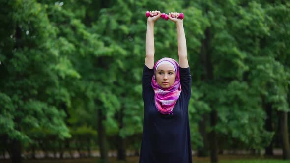 Young Muslim Woman in a Burqa Doing Exercises with Dumbbells in a Summer Park