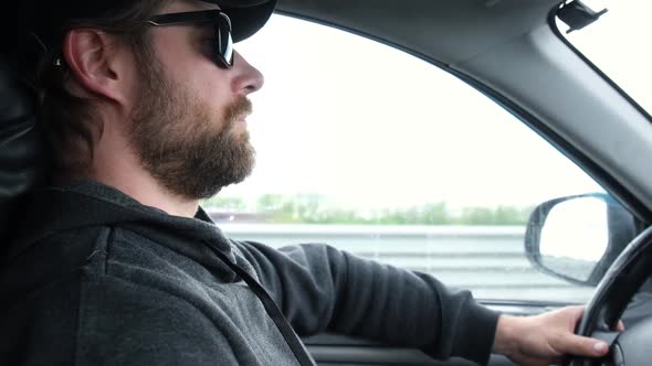 Bearded Man in a Cap and Sunglasses Driving a Car During Road Trip
