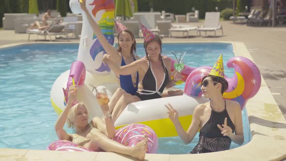 Four Cheerful Young Caucasian Women Dancing in Water Pool on Unicorn and Donut Swimming Rings