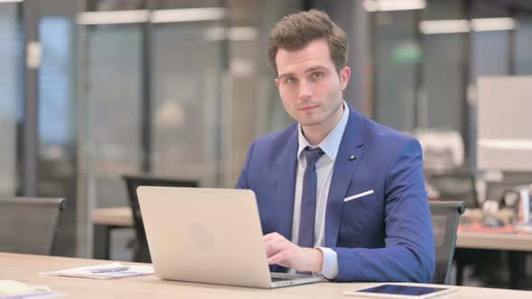 Businessman Pointing at Camera While Using Laptop in Office