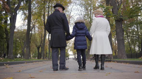 Back View of a Caucasian Senior Couple Walking with a Girl in White Hat Along the Alley