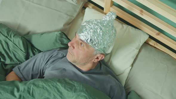 A Man in a Foil Cap Lies in Bed Holding A Cardboard Sign That Says 'FAKE'