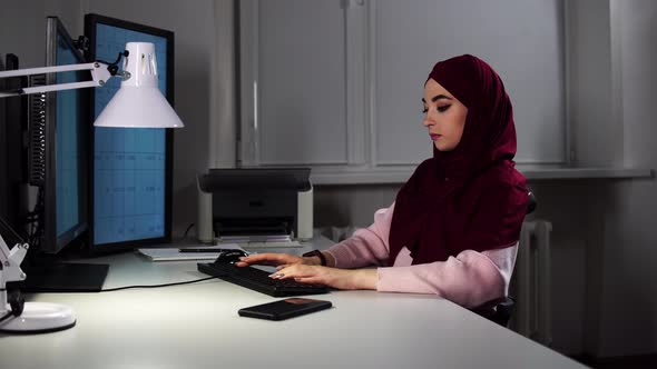 A Young Woman in a Hijab Sits at Desk in Office in Front of Computer and Types on a Keyboard