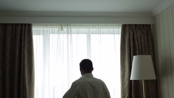 Handsome Man Putting on Shirt Standing Near Window at His Room in Morning.