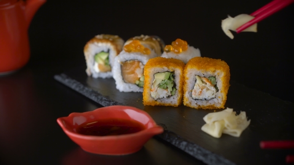 Hand with Red Chopsticks Taking Ginger and Roll Sushi From Dark Plate