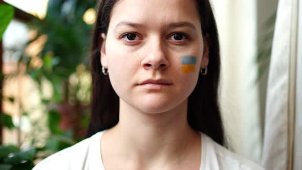 A Sad Young Ukrainian Girl with the Flag of Ukraine on Her Face is Looking to Camera