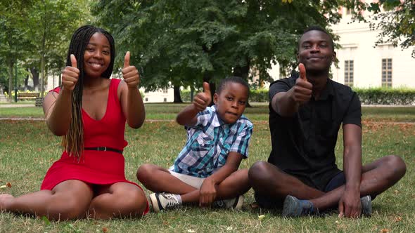 A Black Family Sits on Grass in a Park and Shows Thumbs Up To the Camera with Smiles