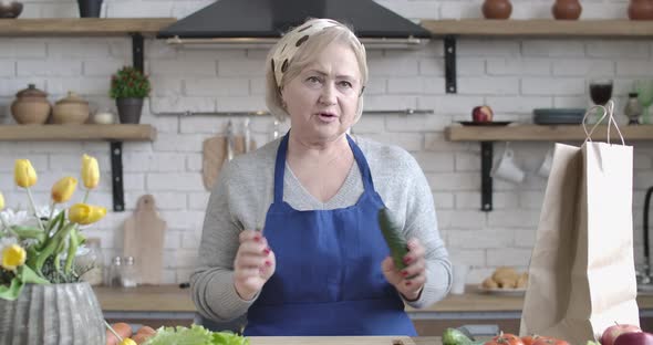 Portrait of Confident Senior Caucasian Woman Talking in Kitchen with Cucumber and Knife in Hands