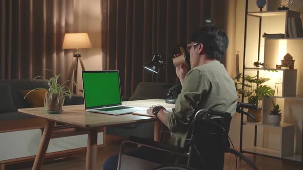 Asian Man Sitting In A Wheelchair While Working On Green Screen Laptop And Drinking Coffee At Home