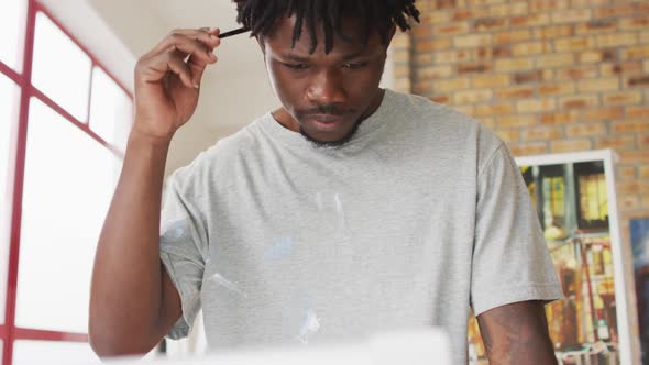 African american male painter holding brush and thinking in artist studio