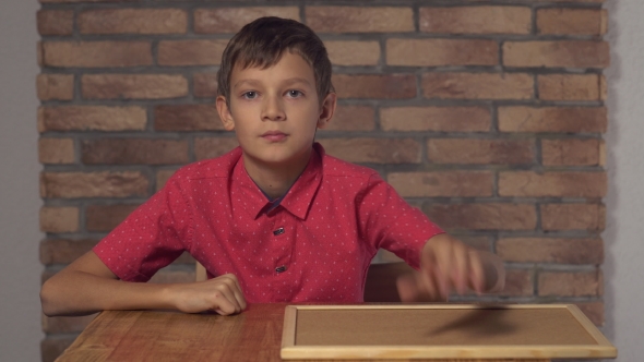 Child Sitting at the Desk Holding Flipchart with Lettering Like on the Background Red Brick Wall.