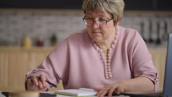 Elderly Woman is Working Remotely From Home Counting By Calculator and Filling Report By Laptop