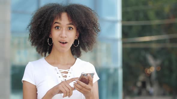 Young Millennial African American Woman Looking in Smartphone, Looking Surprised at Camera Raise