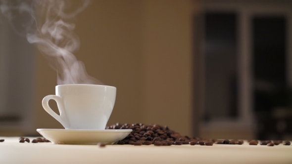 A White Cup of Evaporating Coffee on the Table with Roasted Beans in