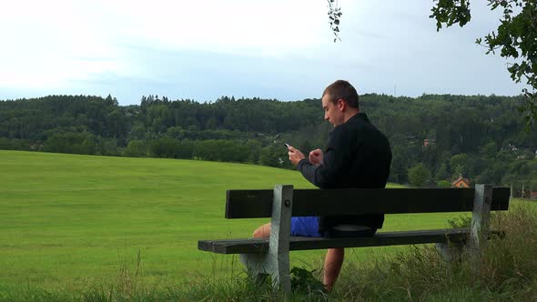 Young Man Sits on the Bench in Nature (Field with Forest) and Works on Smartphone