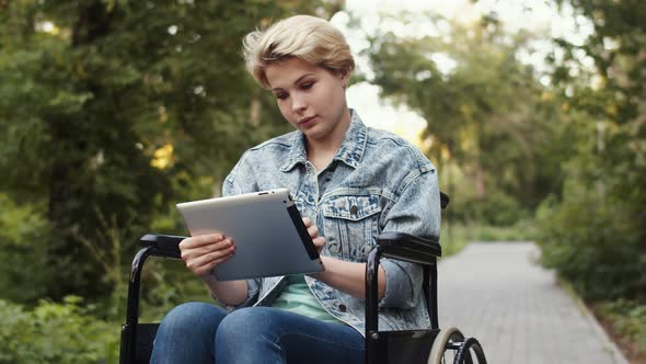 Young Attractive Disabled Woman is Sitting Wheelchair in Park with Tablet in Her Hands and Typing