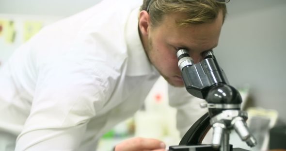 Scientist Is Looking Through Microscope and Writing Data on Tablet. alt