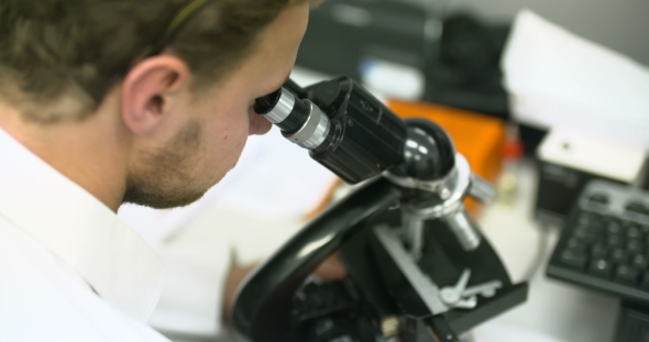Scientist Is Looking Through Microscope and Writing Data on Tablet. alt