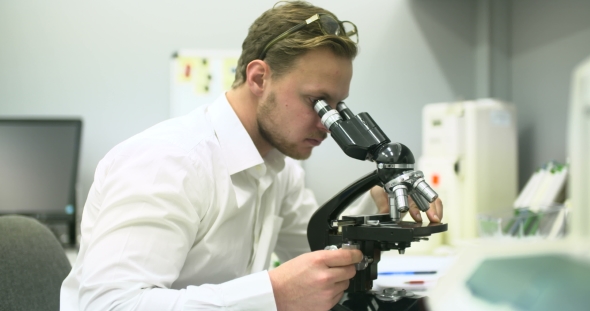 Scientist Is Looking Through Microscope and Writing Data on Tablet.