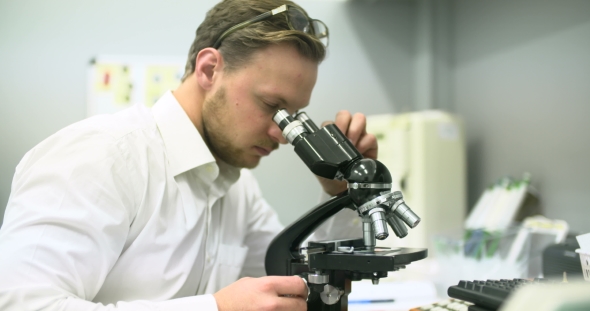 Scientist Is Looking Through Microscope and Writing Data on Tablet.
