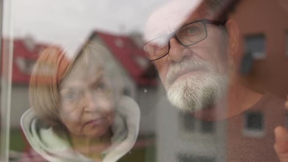 Elderly Married Couple Standing By Window Together Caring Husband Hugging Wife