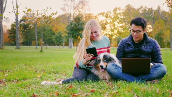 Young People Relaxing in a Park. Asian Man Uses a Laptop, Woman Reading Something on a Tablet