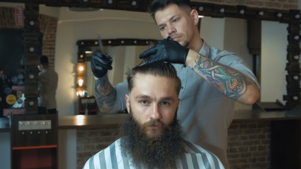 Barber with Dark Hair and Tattoo Wearing Blue Shirt and Black Gloves Doing a Haircut for Client with