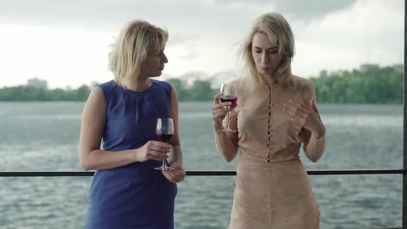 Middle Shot of Beautiful Caucasian Women Standing with Wineglasses on River Bank and Talking
