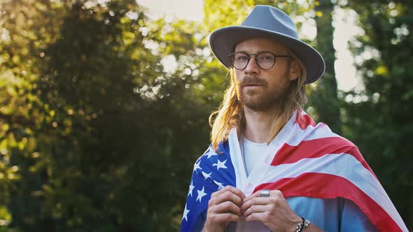 Young Male in Casual Outfit Glasses and Hat is Wrapped in Flag of USA