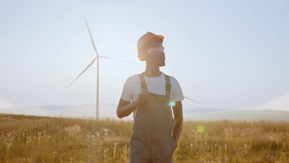 African American Man Standing on Field with Huge Windmills During Summer Sunset