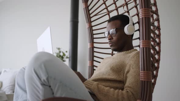 Serious Black Male Freelancer Working on Laptop in Hanging Rocking Chair at Home