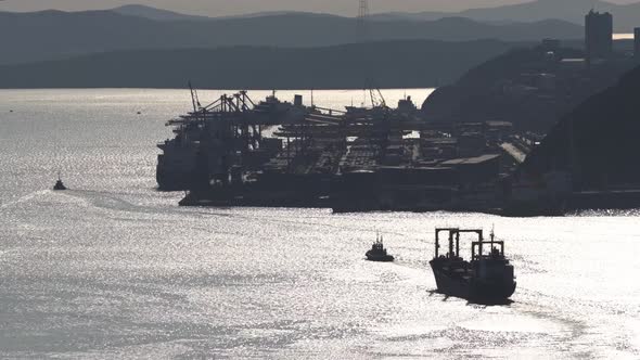 Tug Takes a Large Cargo Ship Out of the Port Into the Open Sea Along the Strait in Silhouette
