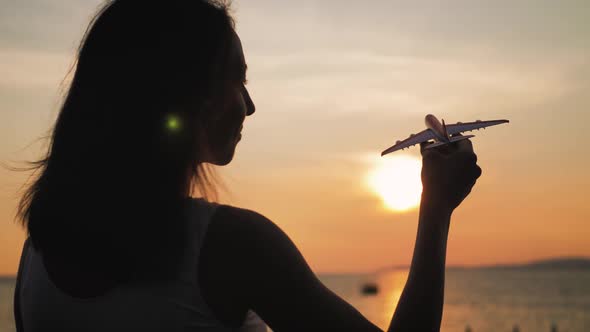 Girl Silhouette Holding Airplane Model in Her Hand During Sunset at Sea. Thinking Idea Concept.