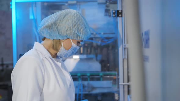 A Woman Operator in a Mask and White Coat Pushes Buttons on the Production Equipment