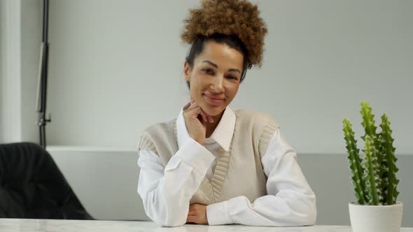 Portrait of a Fashionable Black Curlyhaired Woman with a Beaming Smile Sitting at a Desk in a Modern