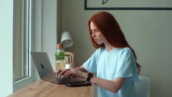 Serious Redhead Young Woman Freelancer Thinking During Working on Laptop Computer Sitting at Table