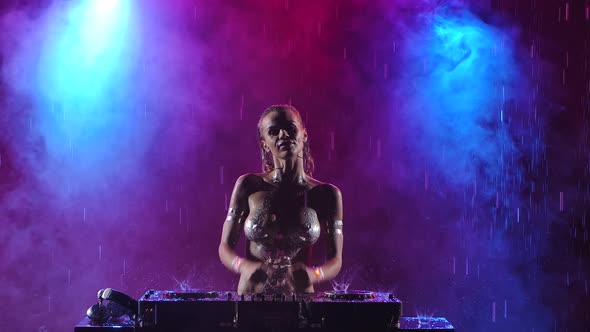 Sexy Female DJ Dancing and Mixing Music on a Console in the Rain. Nude DJ with Silver Bodypainting