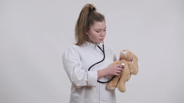 Concentrated Caucasian Girl in Doctor Gown Listening Teddy Bear with Stethoscope and Looking at