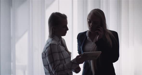 Two Women Discussing a Tablet Display While One Woman Is Also Talking on a Smartphone