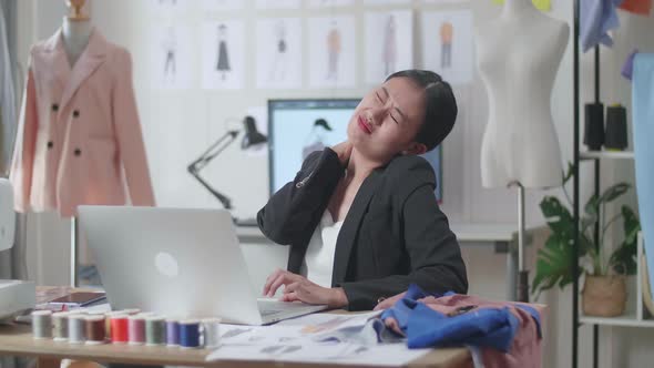 Asian Female Designer In Business Suit Working On A Laptop And Stretching While Designing Clothes