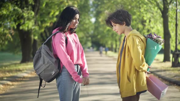 Shy Caucasian Teenage Girl with Backpack Standing on Park Alley As Loving Boy Approaching with Gift