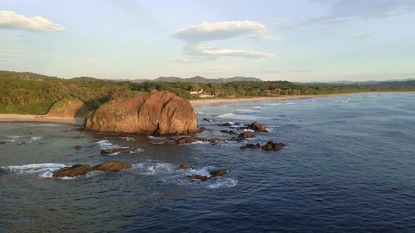 Drone approaching a big beach rock then passing it closely and flying over people enjoying the sunse