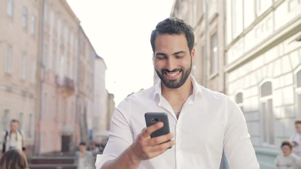 Smiling Man Using Mobile Phone And Walking Along City Street