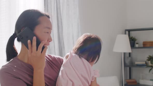 Happy Asian Mom Holding Little Daughter and Speaking on Phone