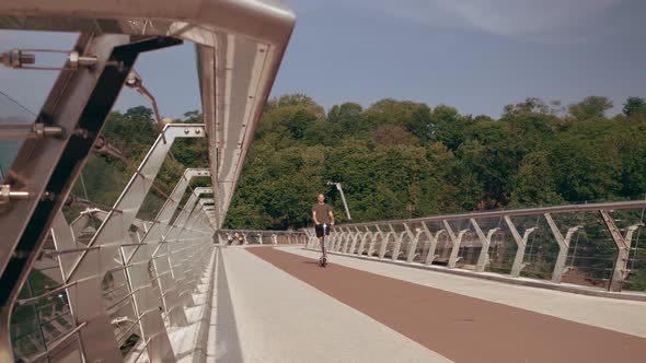 Modern Young Man Rides an Electric Scooter Across the Pedestrian Bridge Overlooking the City