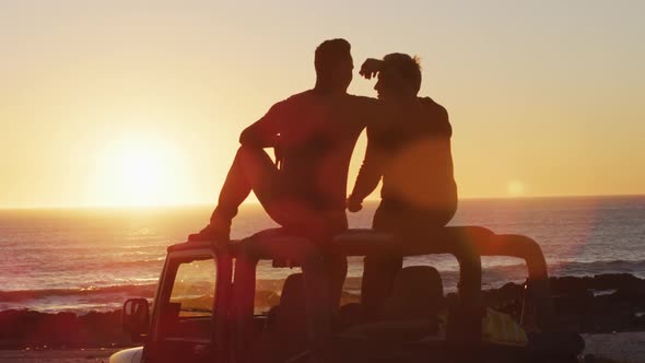 Happy caucasian gay male couple sitting on car roof embracing at sunset on the beach
