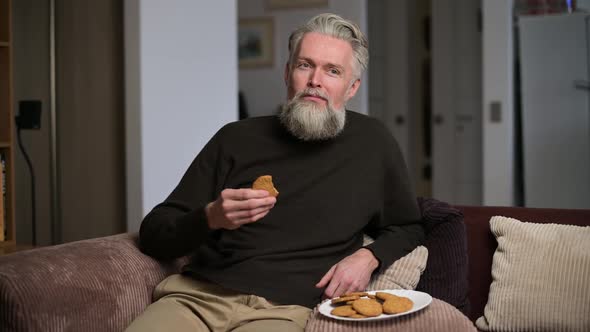 An adult gray-haired man with a beard sits at home on a drink and eats a cookie
