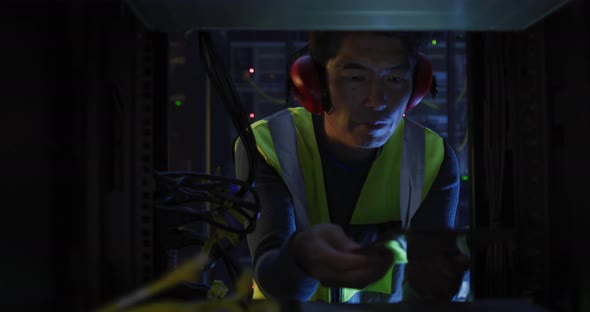 Asian male it technician wearing headphones and using tablet checking computer server